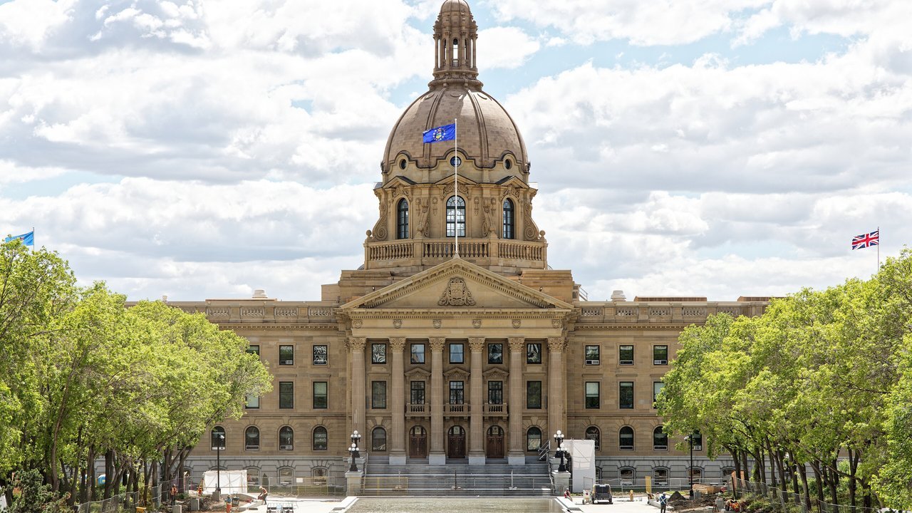 Stand-in, not a photo of this event: the domed sandstone Alberta Legislature Building in Edmonton on a cloudy June 2024 day, with Alberta’s blue flag atop the dome, a Union flag at right, green trees on both sides and construction fencing near the front steps