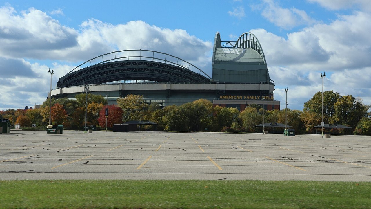 Stand-in, not a photo of this game: American Family Field in Milwaukee in October 2023, its arched grey roof and a sign reading American Family Field above autumn trees, seen across an empty parking lot under a partly cloudy sky