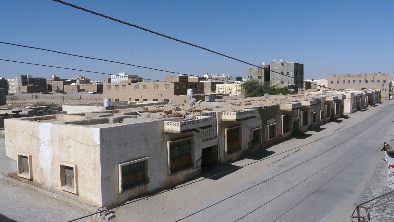 Stand-in, not a photo of this event: December 2009 photo of a street in the old city of Ataq, the Shabwa provincial capital in Yemen, with a row of low pale flat-roofed buildings beside a nearly empty paved road with one person walking at the far right, taller tan buildings behind, power lines overhead and a clear blue sky