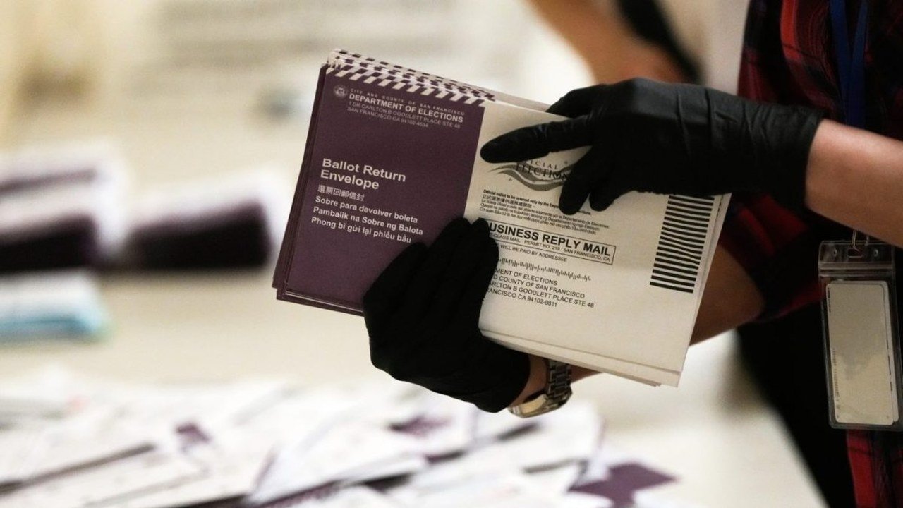 Two hands in black gloves hold a stack of purple and white San Francisco Department of Elections ballot return envelopes over a table covered in more envelopes; the worker’s face does not appear
