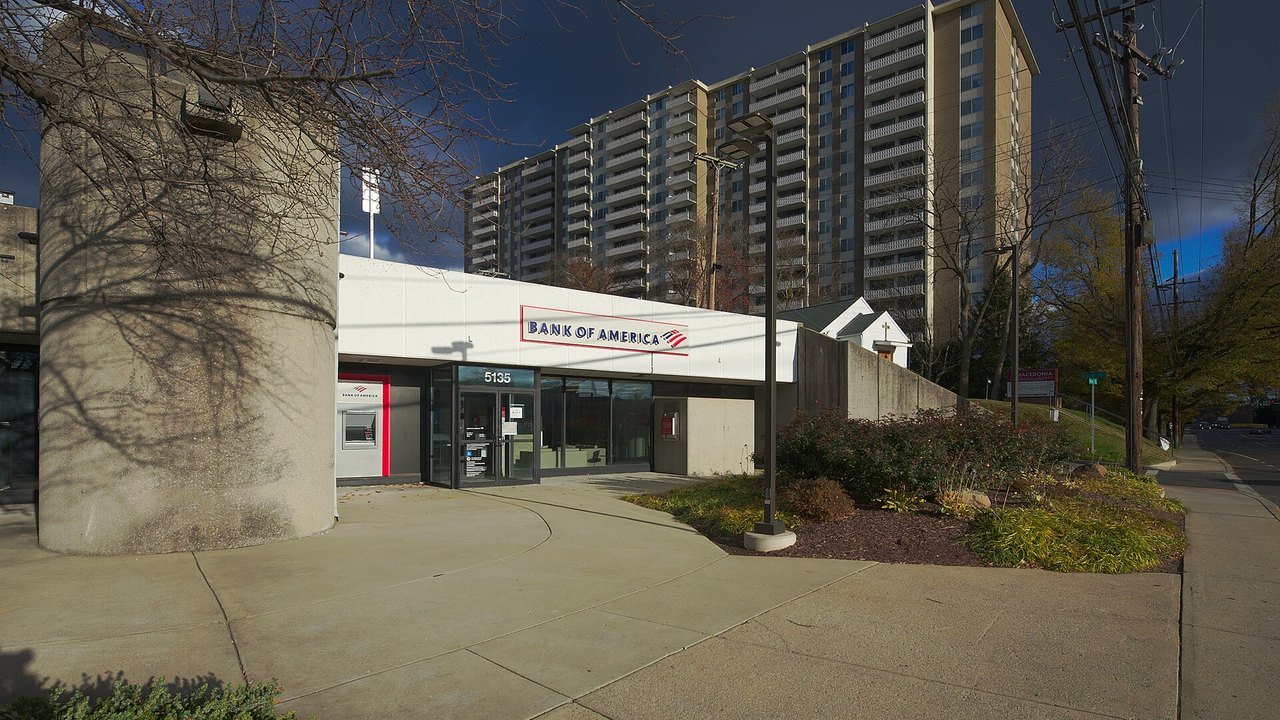 Stand-in, not a photo of this event: a Bank of America branch on River Road in Bethesda, Maryland, with a red and blue Bank of America sign, an ATM, glass doors numbered 5135, a round concrete tower, shrubs and a high-rise apartment block behind it (November 2021)