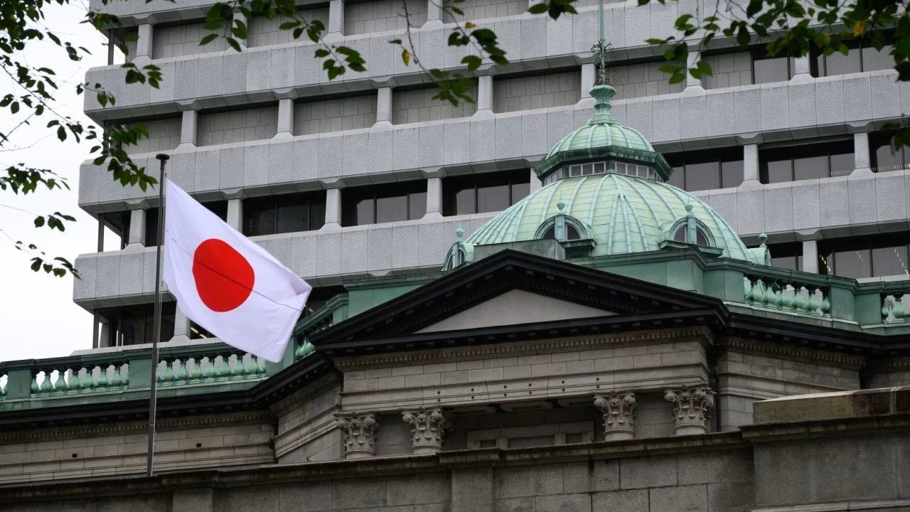 The Japanese flag flies from a pole in front of the green copper dome and stone columns of the Bank of Japan head office in Tokyo, with a grey modern tower behind it, in a photo posted on X