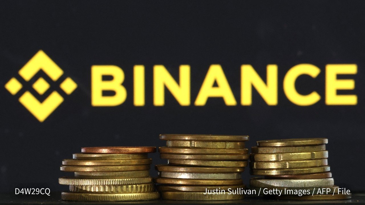 The yellow Binance logo and name glow on a dark screen behind three stacks of gold- and copper-colored coins, in an AFP file photo posted on X
