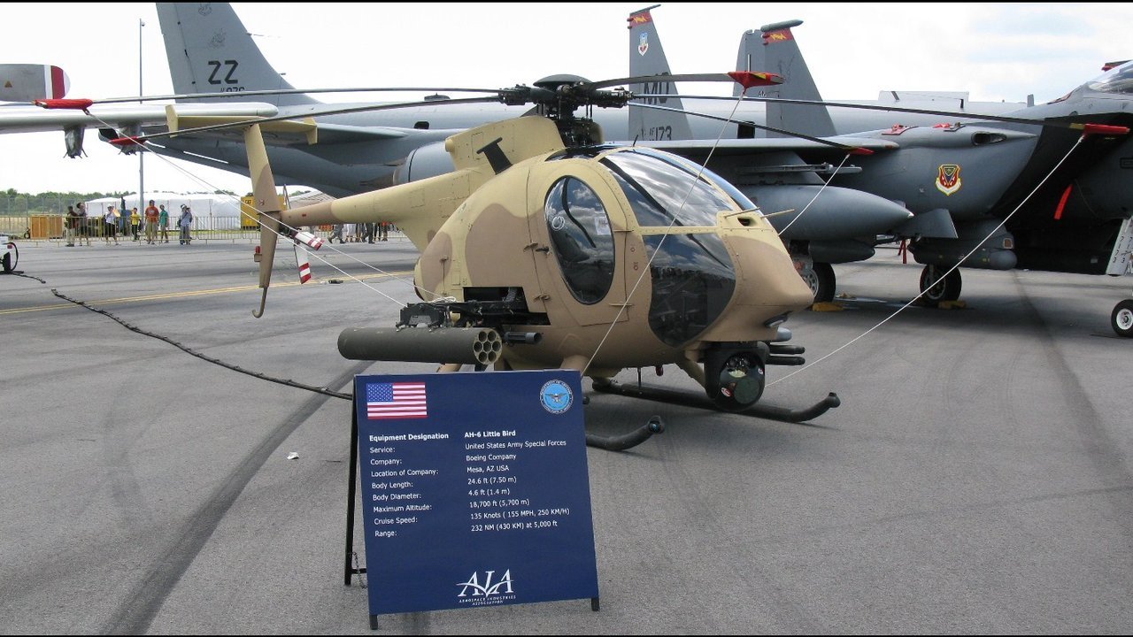 Stand-in, not a photo of this event: February 2010 file photo of a tan-camouflaged Boeing AH-6 Little Bird light attack helicopter with a rocket pod, parked on a static display beside an information sign, with fighter jets behind it