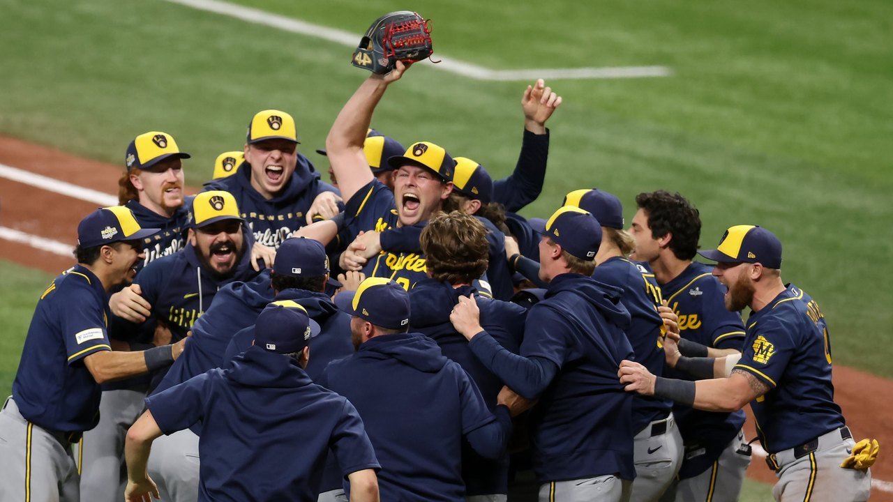 Milwaukee Brewers players in navy hoodies and gold-and-navy caps mob one another on the infield grass at Tropicana Field after the final out of Game 5; one player at center thrusts his glove skyward while teammates shout
