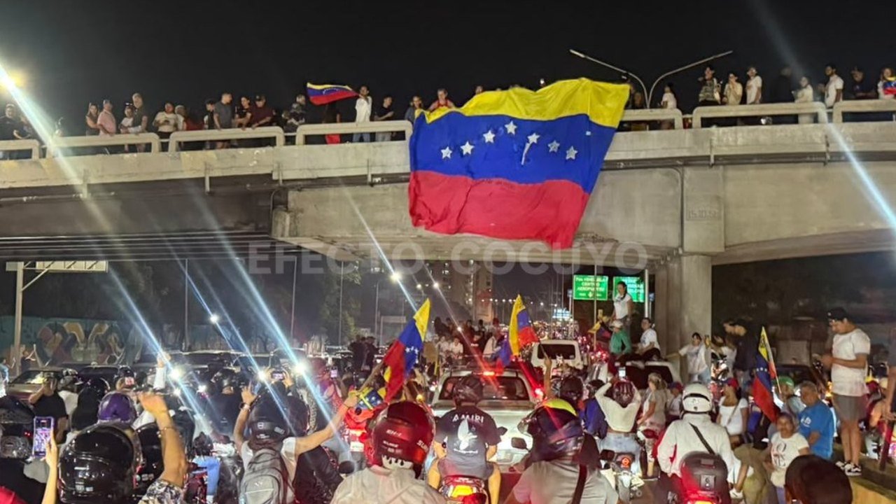 A large Venezuelan flag flies above a night crowd of motorcyclists and cars beneath a highway overpass in Caracas on Sunday, Oct. 25, 2026, as supporters on the bridge watch the caravan welcoming María Corina Machado