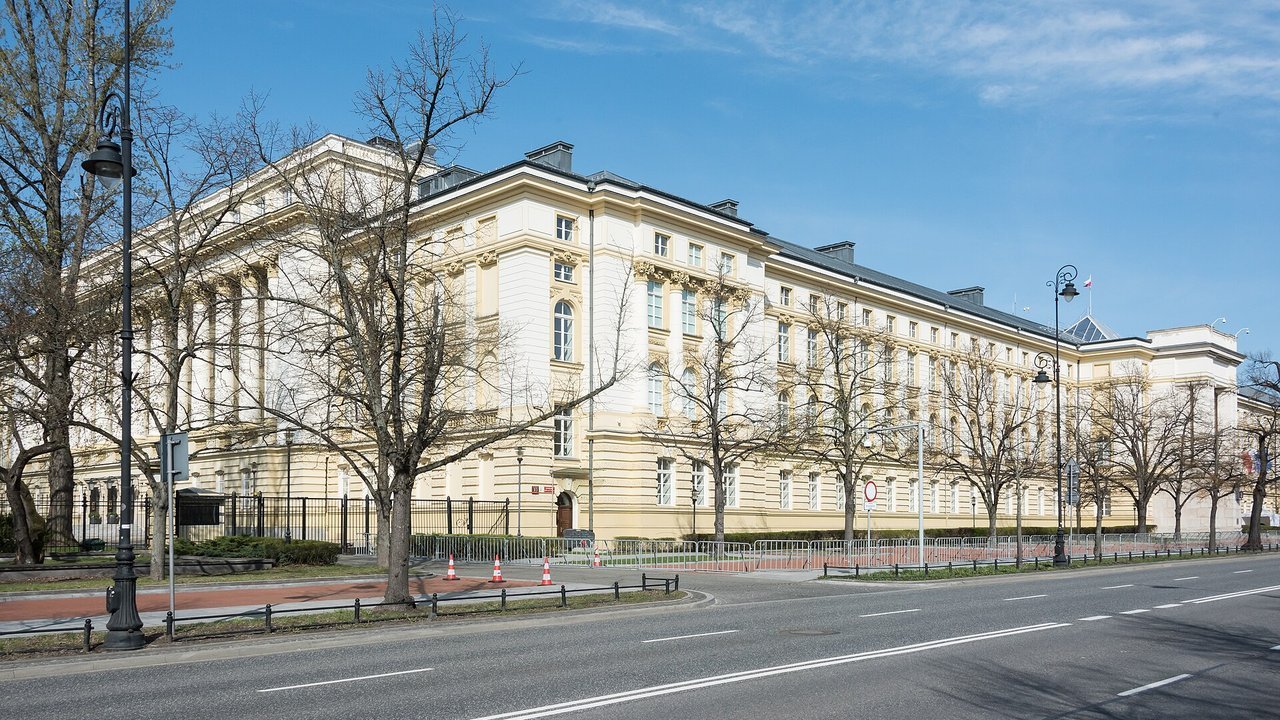 Stand-in, not a photo of this event: the long cream-colored neoclassical building of the Chancellery of the Prime Minister on Aleje Ujazdowskie in Warsaw, behind bare spring trees and an empty street under a blue sky, 2022