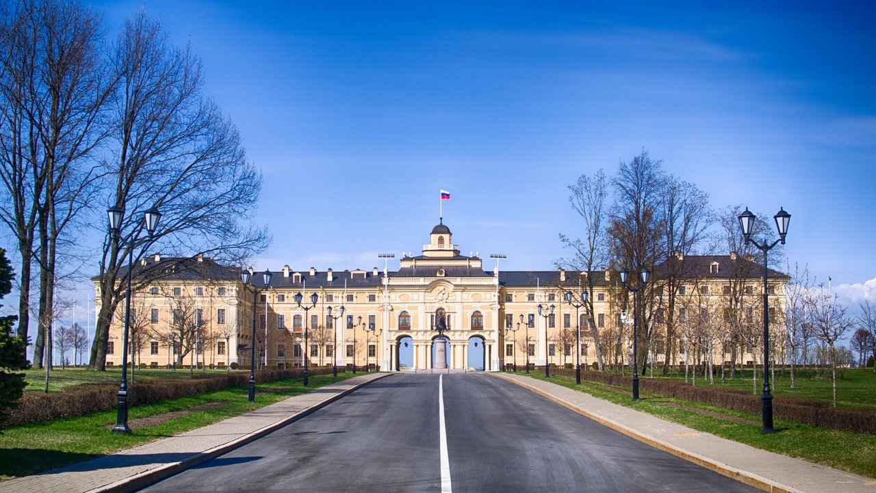Stand-in, not a photo of this event: a long cream-colored palace with a central clock tower flying a Russian flag, at the end of a straight paved road lined with black lampposts, bare trees and green lawns, under a blue sky, in a 2014 photo of the Constantine Palace in Strelna near St. Petersburg