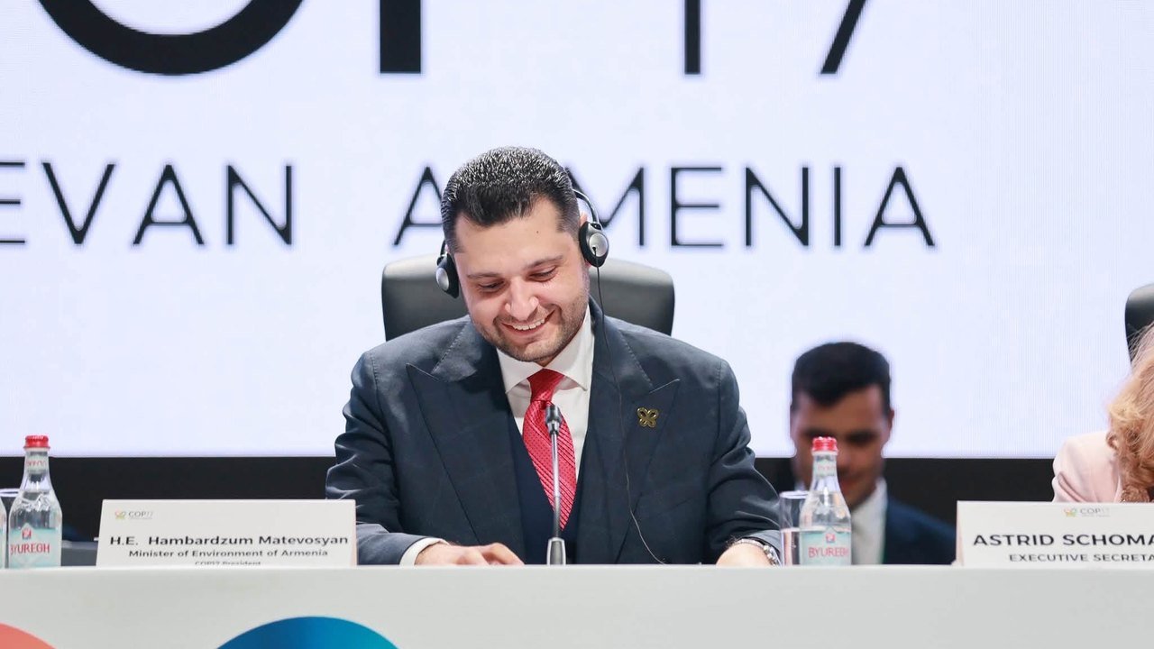 COP17 President Hambardzum Matevosyan, smiling and wearing headphones and a red tie, sits at the dais behind nameplates under a COP17 Yerevan Armenia backdrop