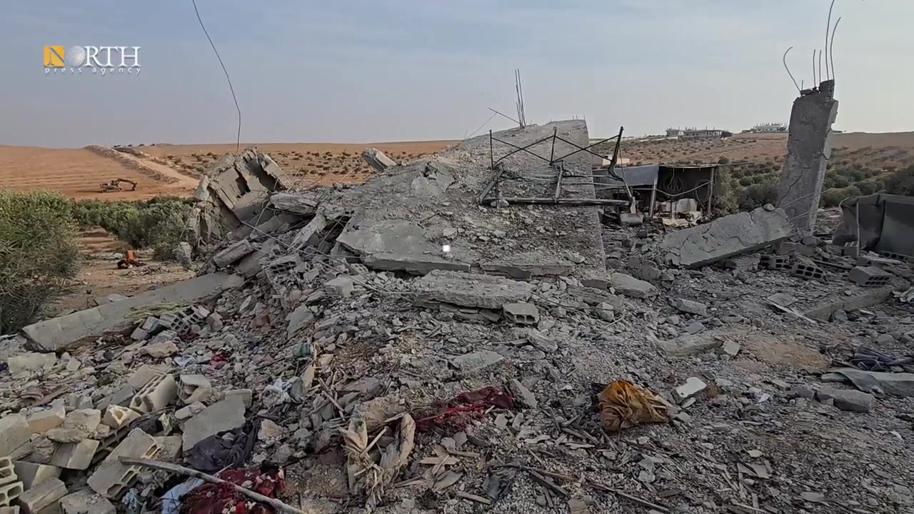 Video still posted on X by @NPA_English: the flattened concrete remains of a house in Daraa’s countryside in southern Syria, with rubble, twisted rebar and scattered fabric in the foreground and farmland and a digger in the distance under a pale sky