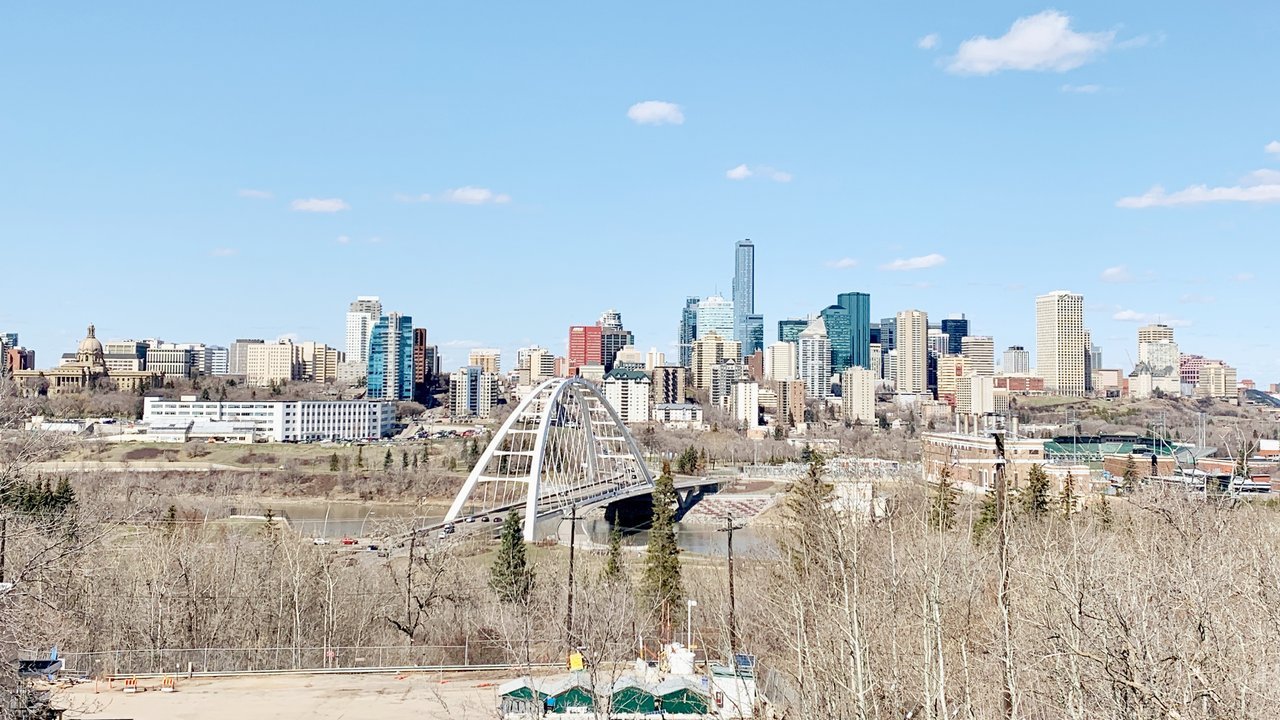 Stand-in, not a photo of this event: downtown Edmonton towers under a blue sky in April 2023, seen from Saskatchewan Drive, with the white arch of the Walterdale Bridge over the North Saskatchewan River and the domed Alberta Legislature Building at far left behind bare trees