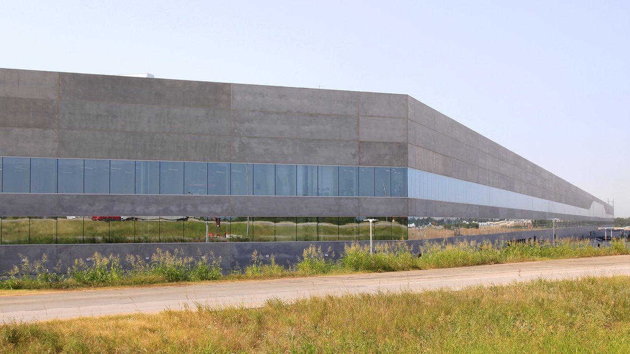 Stand-in, not a photo of this event: the long gray concrete wall of Tesla’s Gigafactory Texas Building 1 near Austin in June 2022, with two bands of reflective glass windows, tall green and yellow grass along a paved road in front and a pale blue sky above