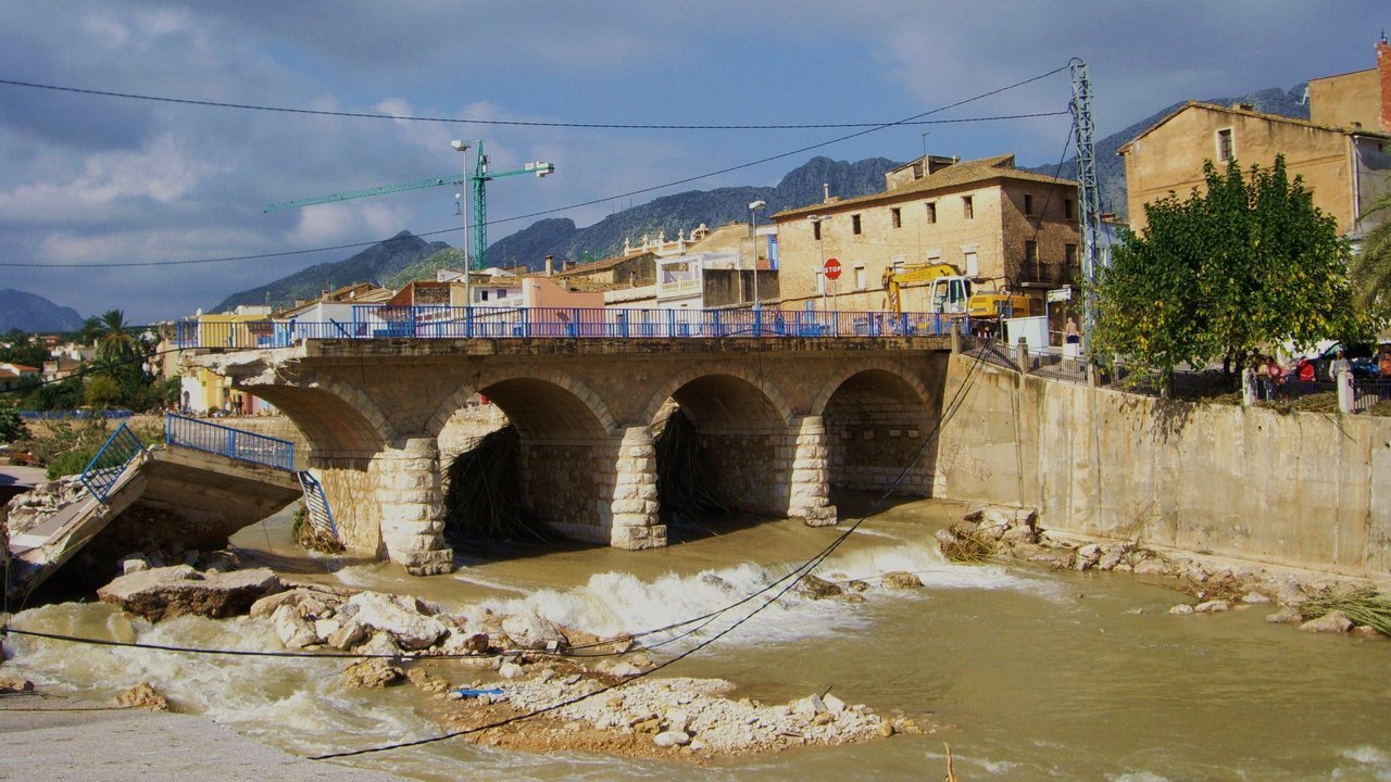 Stand-in, not a photo of this event: October 2007 photo of the Girona river at Beniarbeig in Spain’s Marina Alta after an earlier flood, with brown water rushing under a stone arch bridge, a broken footbridge section on the left, rocks in the riverbed, and houses and mountains behind under a cloudy sky