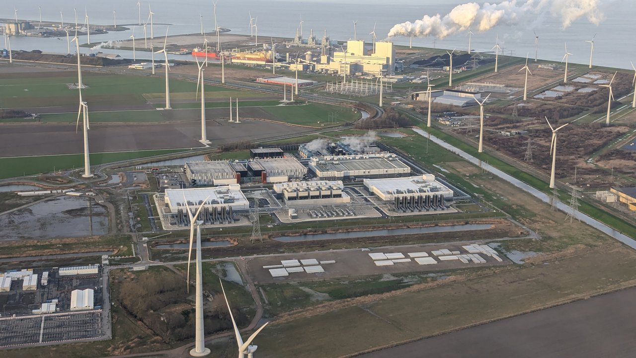 Stand-in, not a photo of this event: aerial view of Google’s data center at Eemshaven in the Netherlands (not the London facility affected by the outage) in January 2023, low white buildings with rows of cooling units surrounded by wind turbines, canals and flat fields, with a power plant releasing white plumes near the sea behind