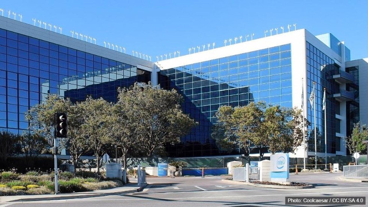 A glass-fronted, multistory office building with white trim under a clear blue sky, trees and a traffic light in front, and a small white Intel sign by the entrance drive