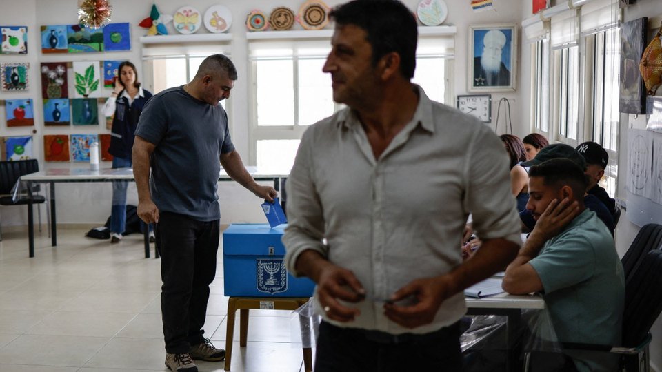 AFP photo posted on X by @Channel8English: a man drops an envelope into a blue ballot box at a classroom polling station in Israel as a polling official in a gray shirt walks past, Oct. 27, 2026