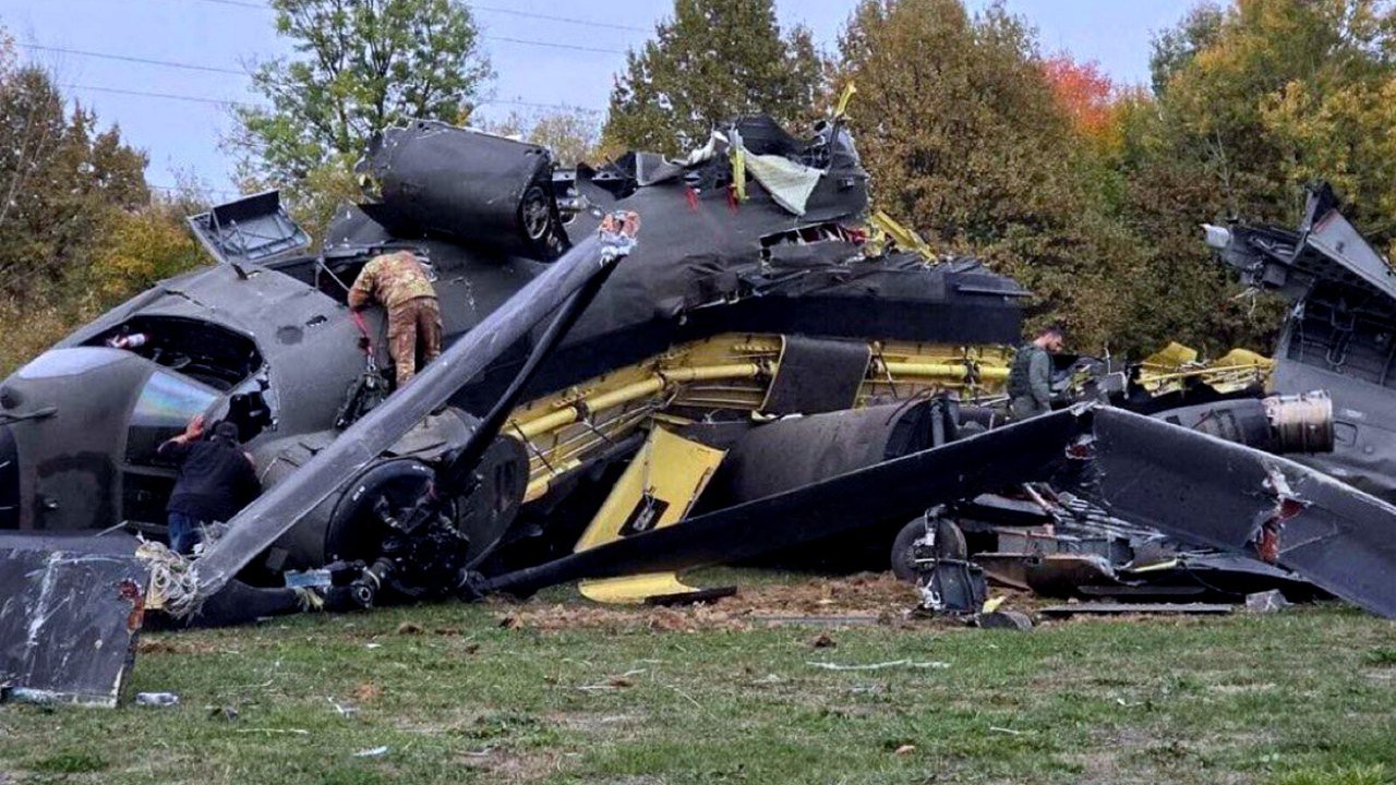 The broken fuselage and rotor blades of a dark green military transport helicopter lie on grass in front of autumn trees, with two people in uniform climbing over the wreckage, in a photo posted on X near Brčko, Bosnia and Herzegovina