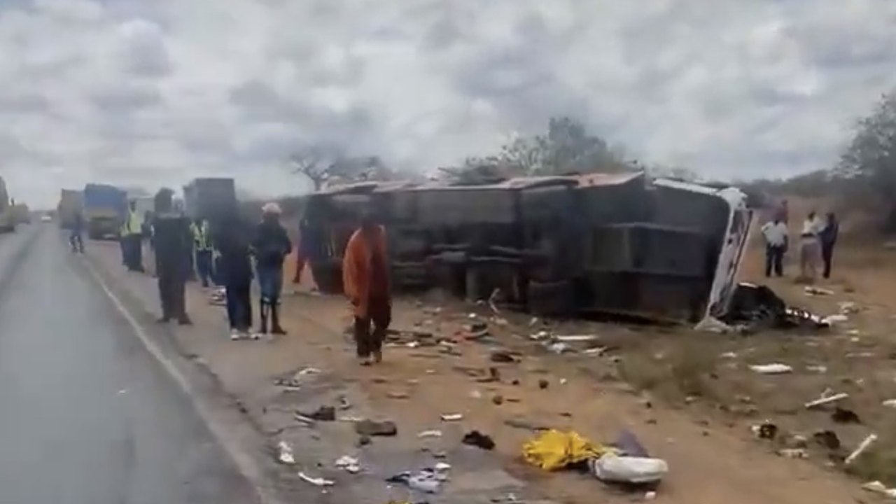 An overturned bus lies on its side in red dirt beside a two-lane highway under a grey sky, with clothing and debris scattered in the foreground, onlookers and officers in reflective vests standing by the road, and lorries queued in the distance, Kanga, near Mtito Andei, Kenya, Oct. 25, 2026
