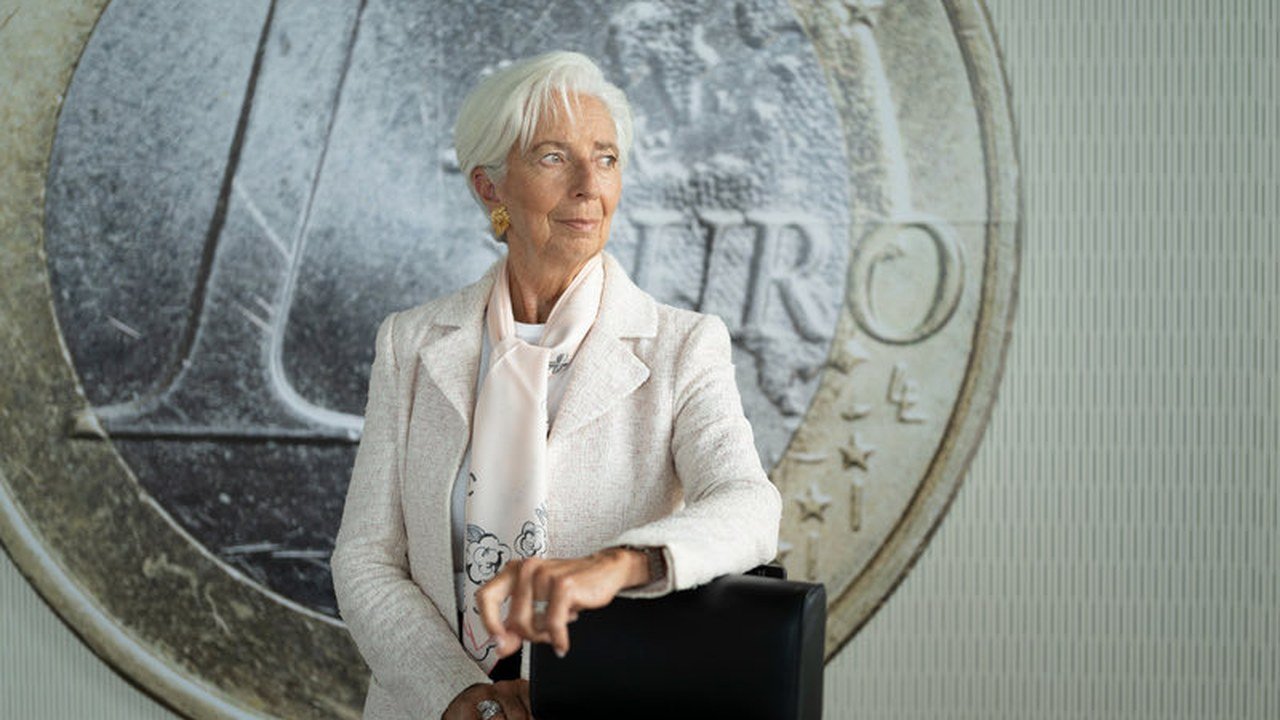 Christine Lagarde, with white hair, a cream jacket and a pale pink scarf, rests one hand on the back of a black chair in front of a large photo of a one-euro coin, in a portrait the European Central Bank posted on X