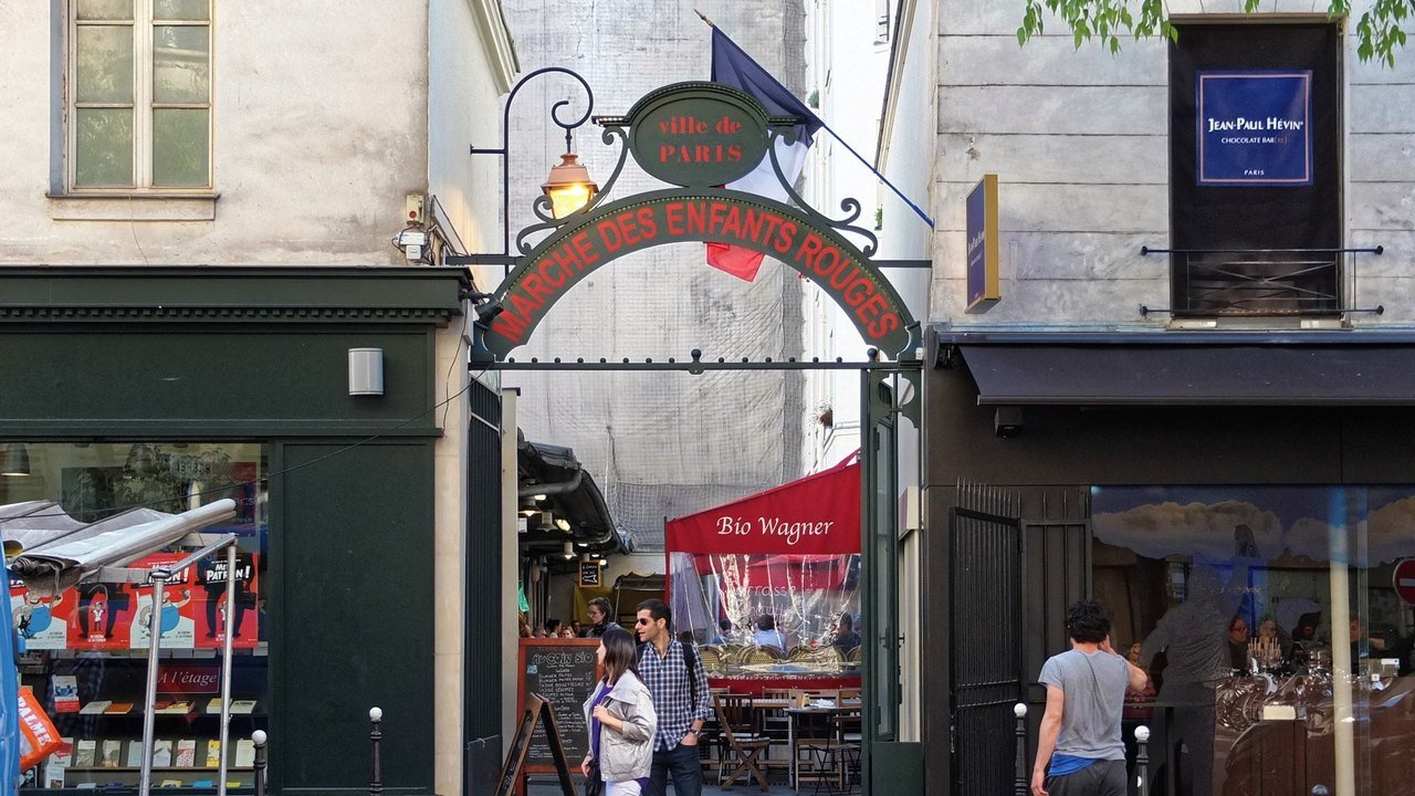 Stand-in, not a photo of the fire: a 2016 file photo of the green iron entrance arch of the Marché des Enfants Rouges in Paris, with red lettering beneath a Ville de Paris sign and a French flag, a red Bio Wagner awning inside and passers-by in the street