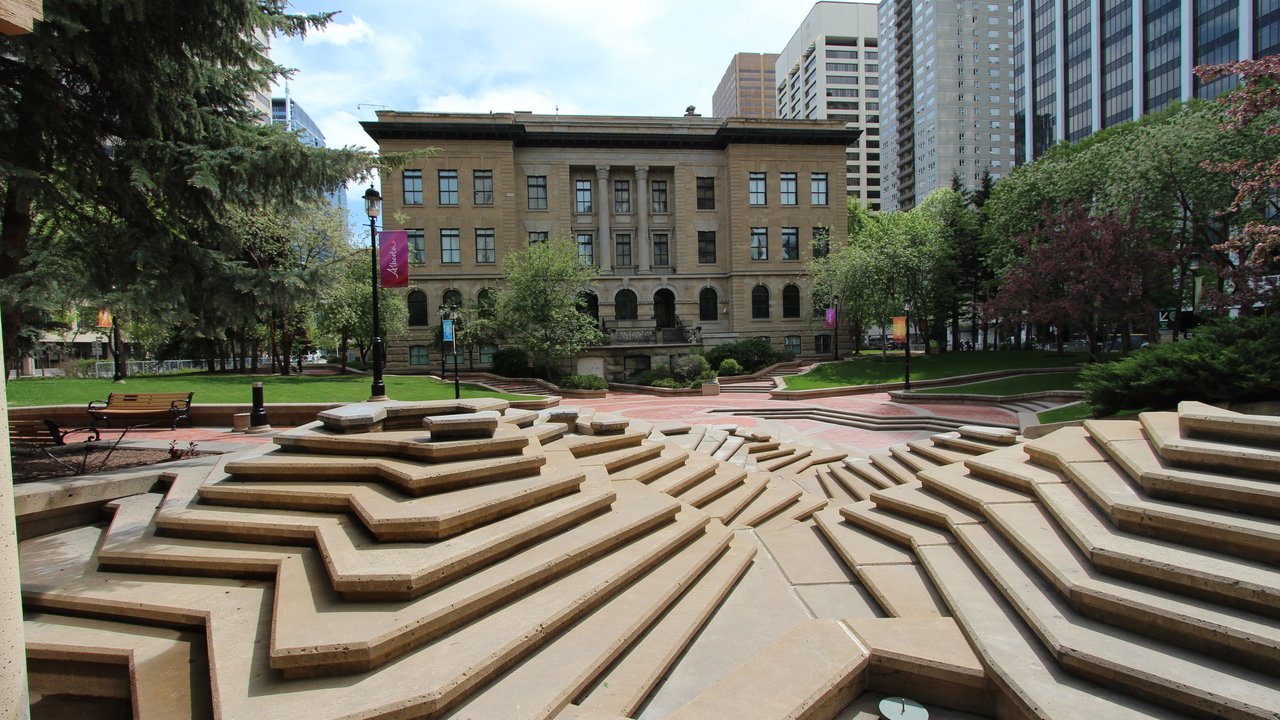 Stand-in, not a photo of this event: a sandstone three-storey building with tall windows behind a lawn and a plaza of tiered, angular stone steps, with downtown Calgary towers behind it, in a 2013 photo its uploader titled the McDougall Centre, Calgary