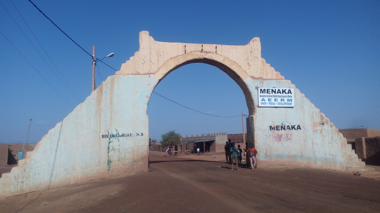 Stand-in, not a photo of this event: a pale, stepped earthen arch over a dirt road, with signs bearing the town’s name, a few people standing in its shade and low buildings beyond, in a 2017 photo of the town of Ménaka, Mali
