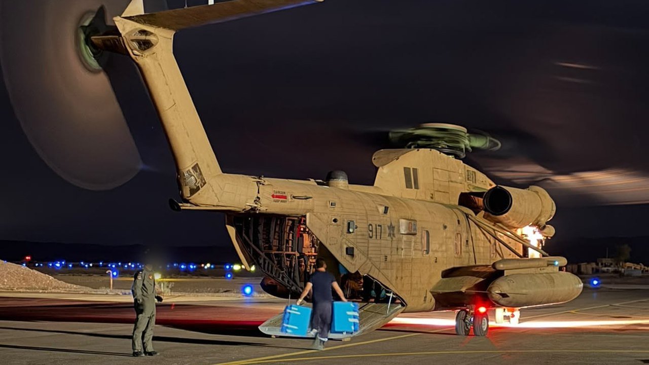 At night on an airfield, a man carries two blue ballot boxes up the rear ramp of a tan Israeli Air Force heavy-lift helicopter with spinning rotors, while a crew member in flight gear stands on the tarmac