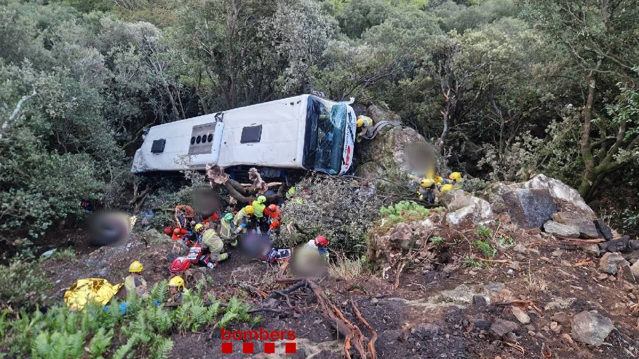 A white bus lies on its side among trees and boulders on a steep, wooded slope as firefighters in yellow helmets and rescuers in orange tend to injured people on the ground below it, with a gold thermal blanket in the foreground, Fogars de Montclús, Catalonia, Spain, Oct. 25, 2026