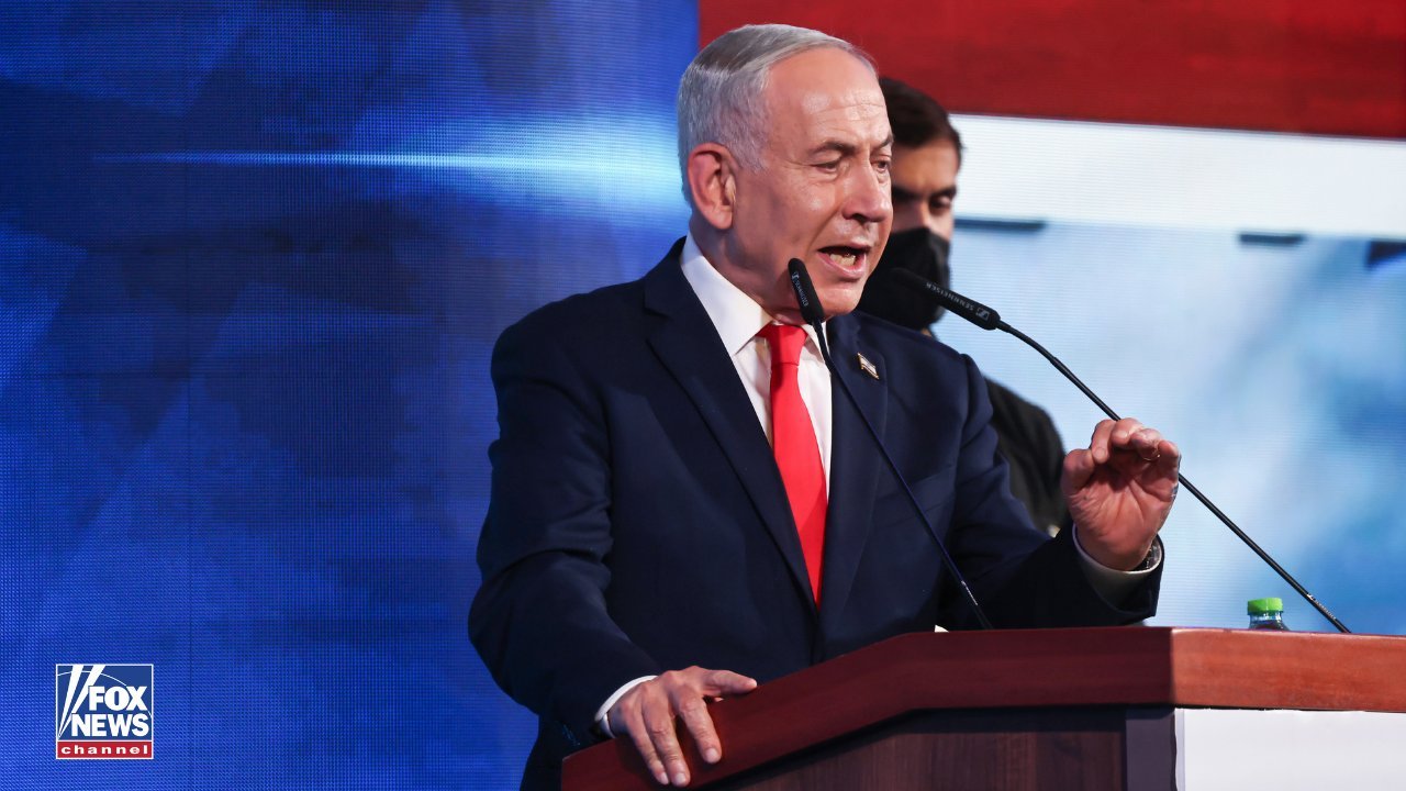 Benjamin Netanyahu in a dark suit and red tie speaks at a wooden podium with a microphone before a blue-and-red stage backdrop, a masked security guard behind his left shoulder