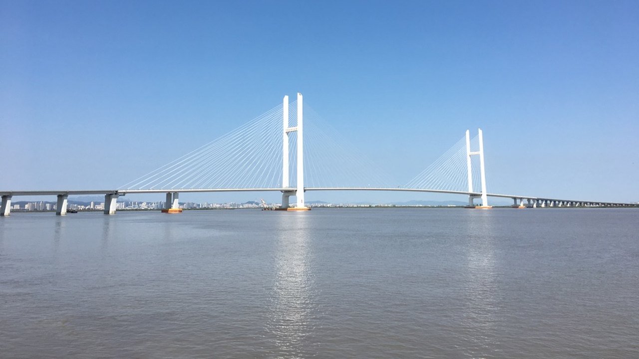 Stand-in, not a photo of this event: a white cable-stayed bridge with two tall H-shaped pylons stretches across a wide, calm river under a clear blue sky, with a low city skyline on the far bank, photographed in 2016