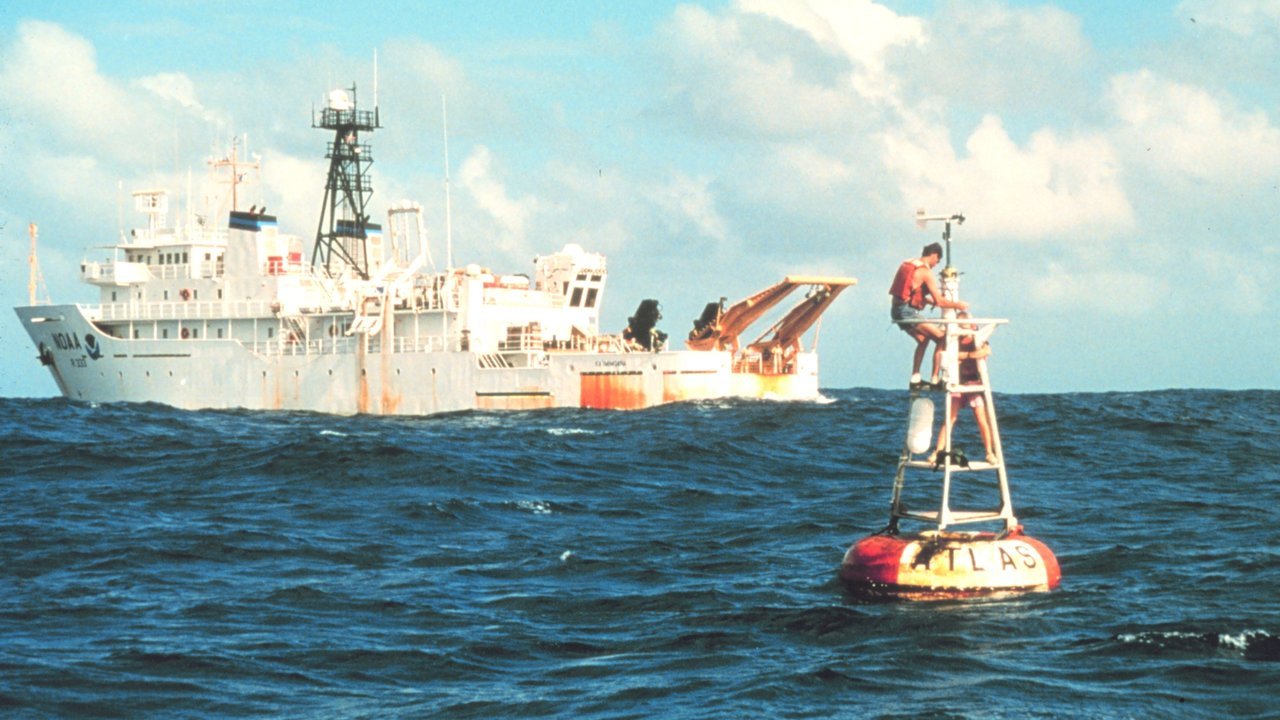 Stand-in, not a photo of this event: an archival NOAA file photo from 1996 or later of the white research ship Ka’imimoana on the open sea, with a crew member standing on the frame of an orange-and-white ATLAS buoy in the foreground