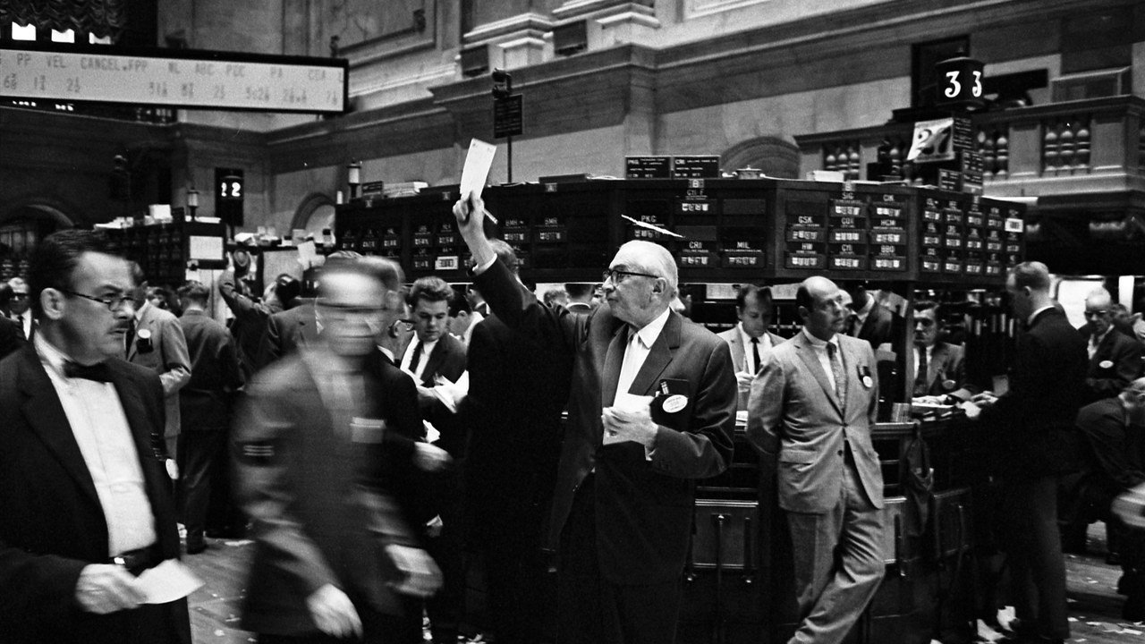 Stand-in, not a photo of this event: crowded New York Stock Exchange trading floor with traders, papers and boards under bright lights, photographed in 1963