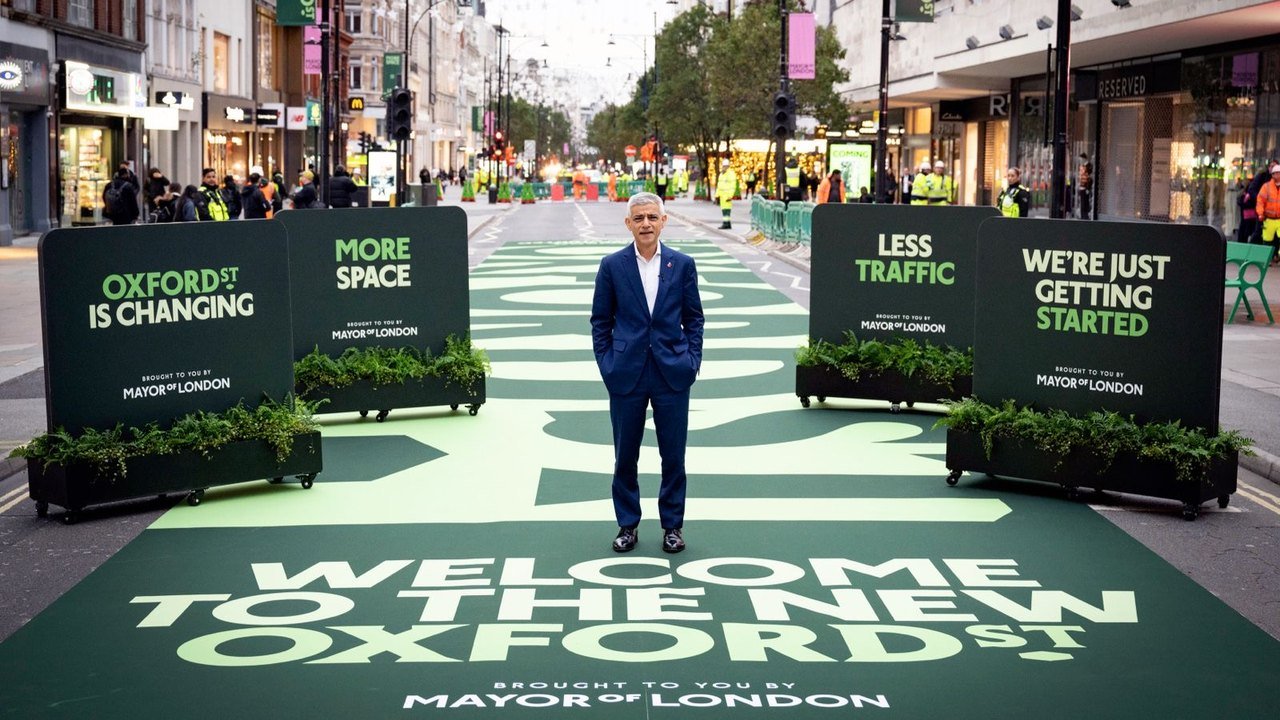 A man in a dark blue suit stands alone on a green painted road surface reading Welcome to the new Oxford St, between planters and signs reading More space and Less traffic, with workers in high-visibility jackets and shoppers behind him, Oxford Street, London, Oct. 26, 2026