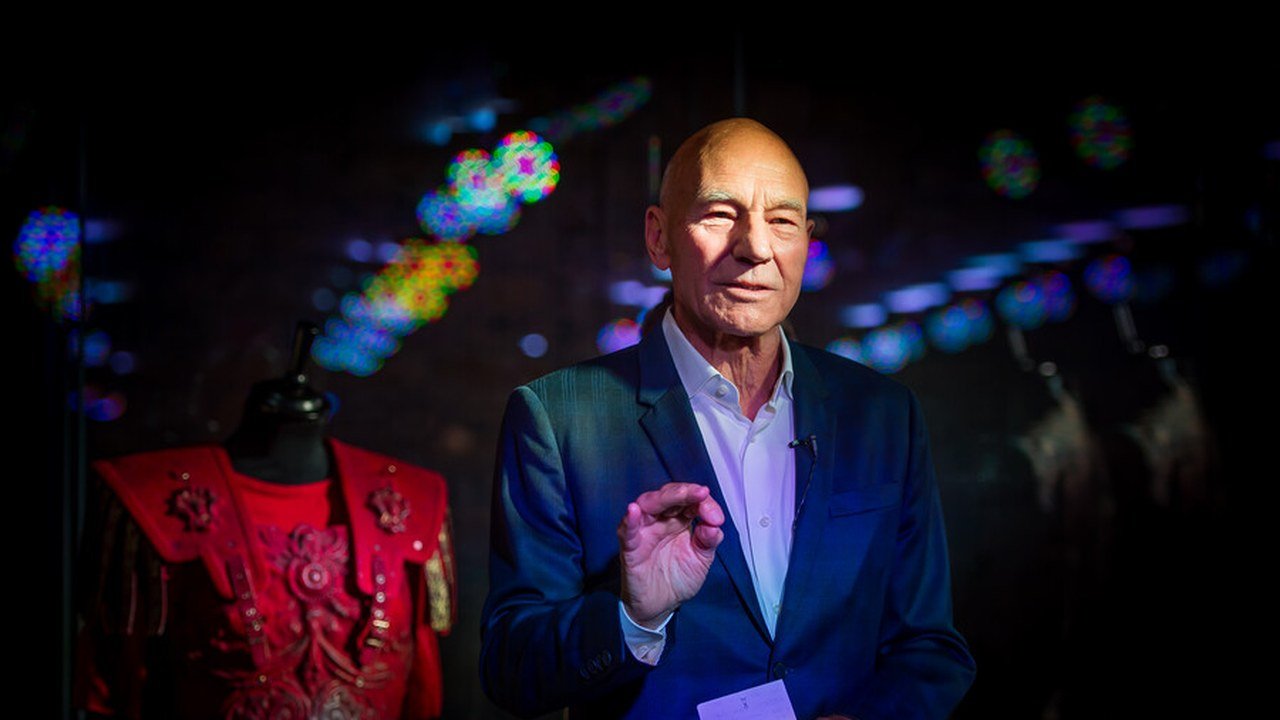 Patrick Stewart, bald and wearing a dark blue suit and white open-collared shirt, gestures as he speaks beside a red embroidered costume on a mannequin, against a dark background with colored lights, in a photo the Royal Shakespeare Company shared on X