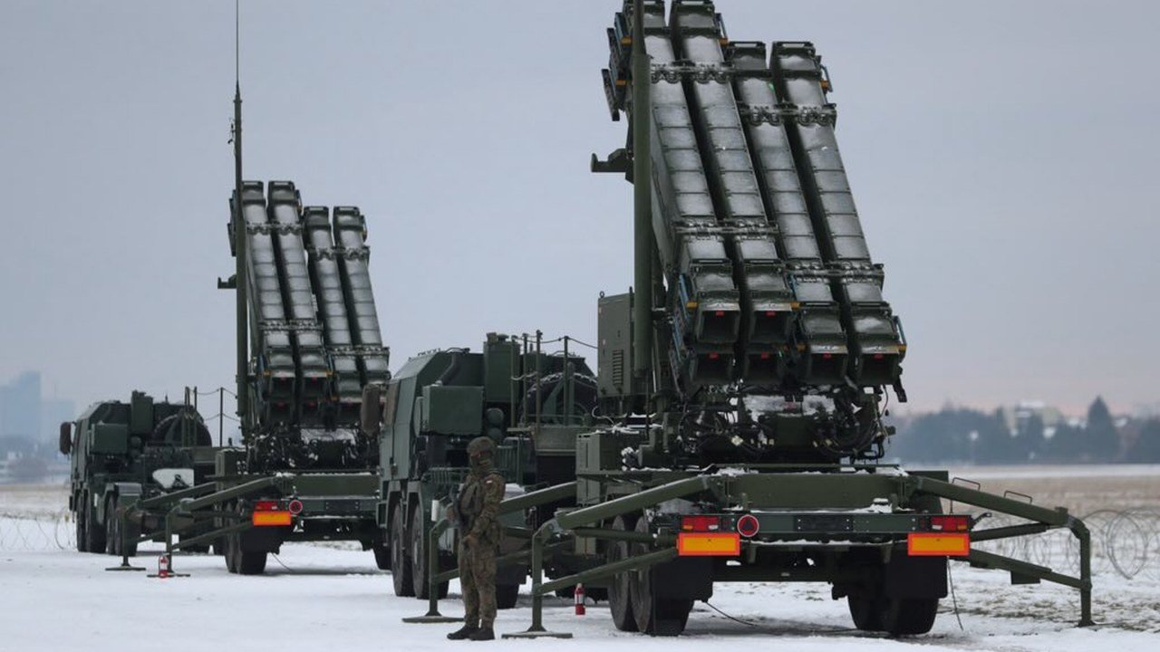 Three Patriot air defense launcher trucks with raised missile canisters stand on snowy ground under a grey sky, with one soldier in camouflage standing beside the nearest truck; no face shows clearly