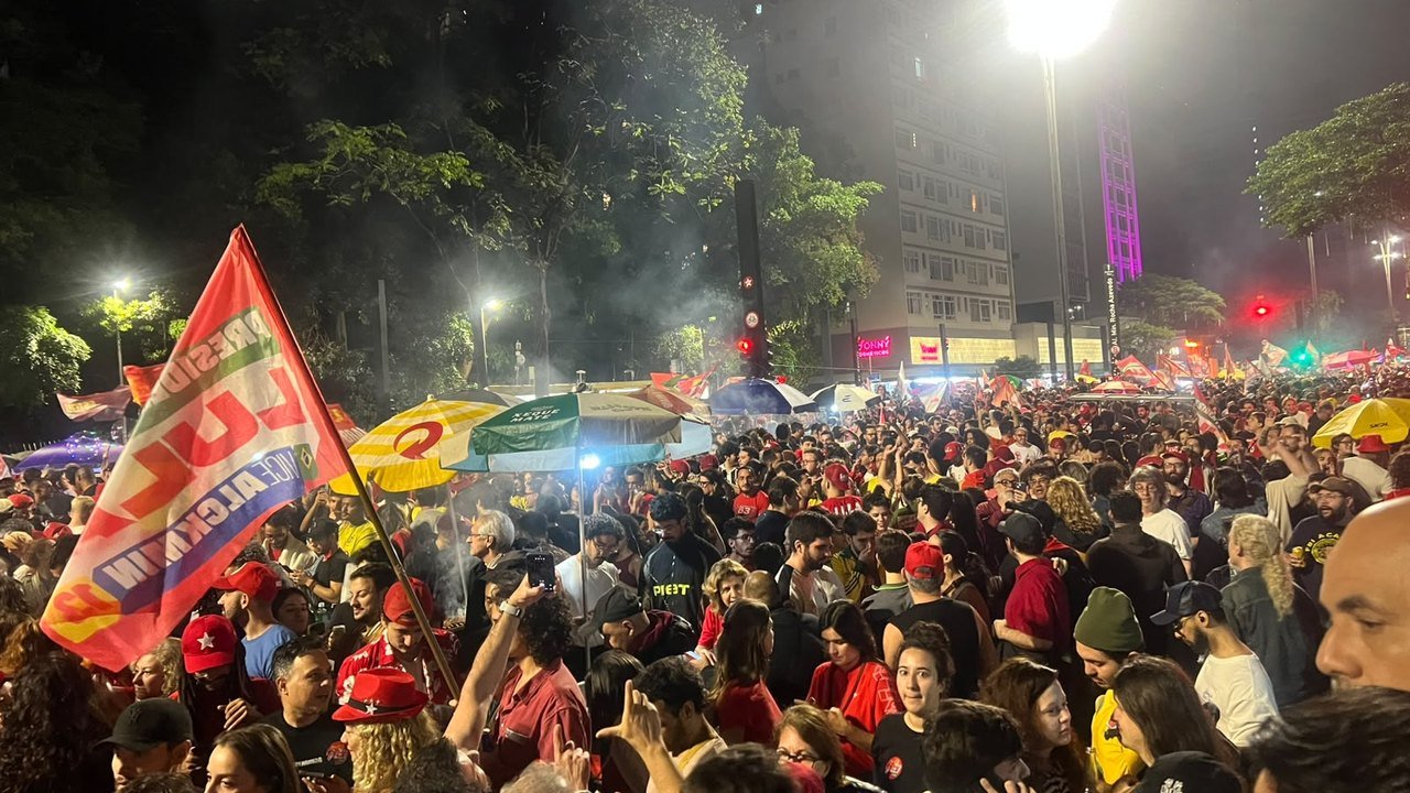 A night crowd fills Avenida Paulista in São Paulo on Sunday, Oct. 25, 2026, after Lula’s runoff win; a red Lula–Alckmin campaign flag waves at left beside striped umbrellas and a tall streetlight