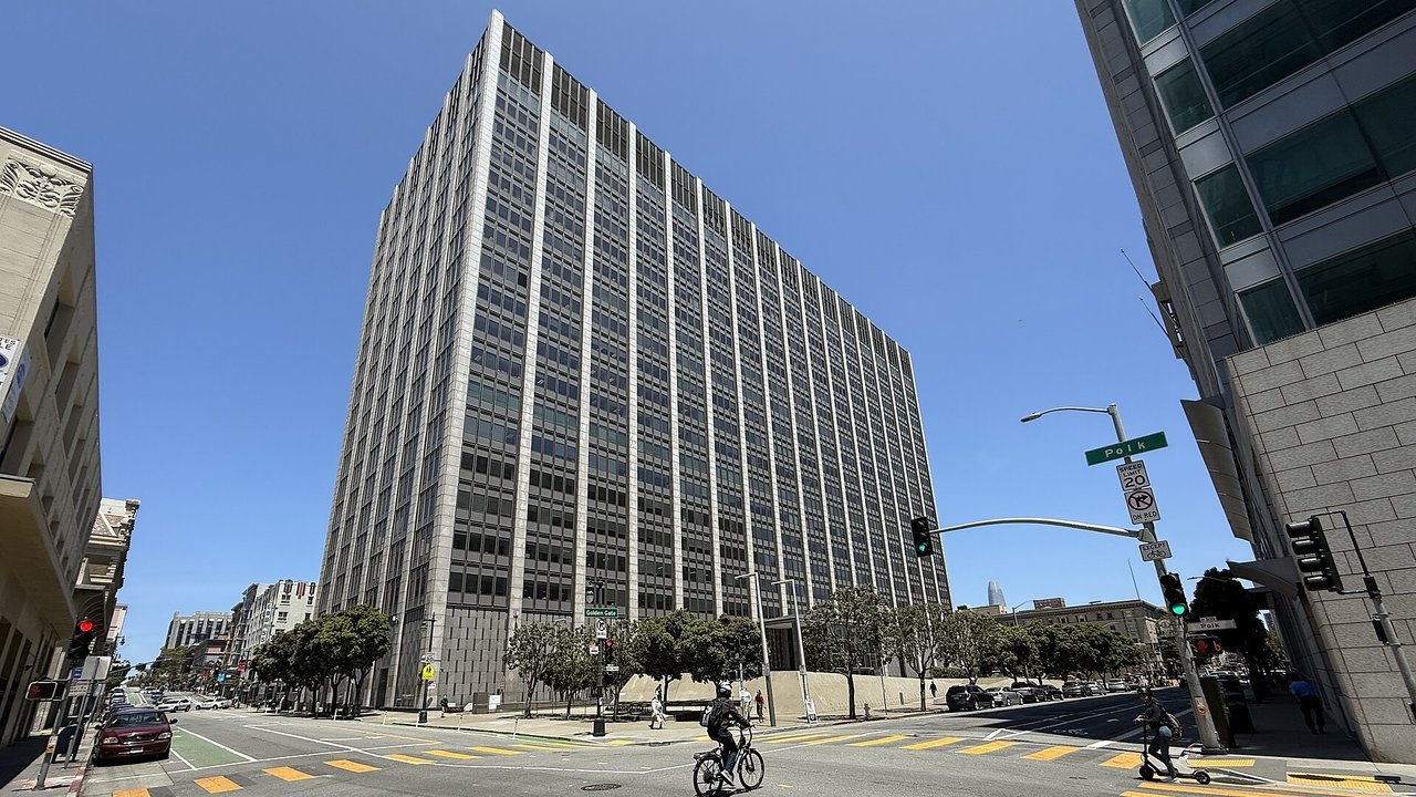 Stand-in, not a photo of this event: the Phillip Burton Federal Building and United States Courthouse in San Francisco, a grey high-rise tower seen from a street crossing with a cyclist in the foreground (June 2025)