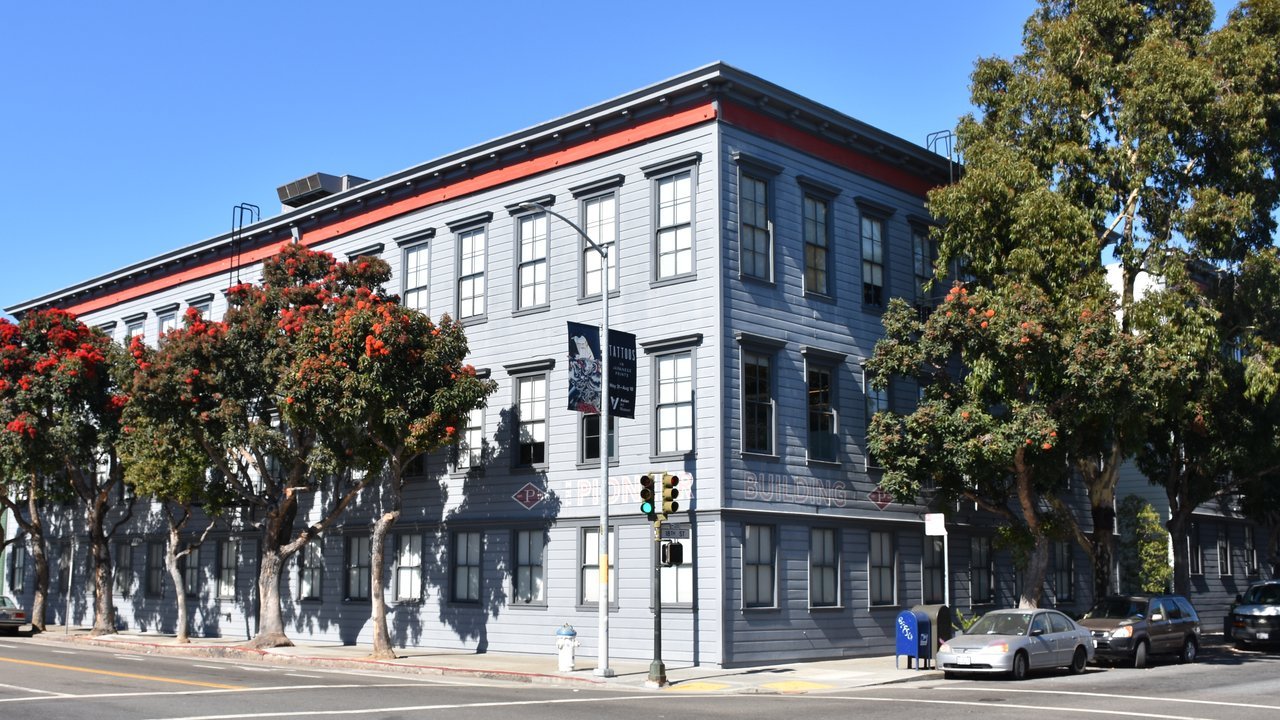 Stand-in, not a photo of this event: a July 2019 file photo of the Pioneer Building, a three-story gray clapboard building in San Francisco’s Mission District that then housed OpenAI offices, under a blue sky with flowering street trees