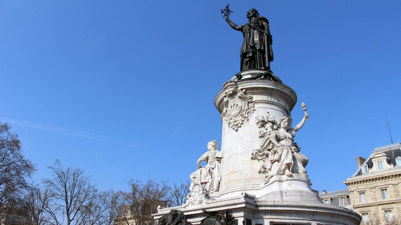 Stand-in, not a photo of this event: a bronze statue of Marianne holding up an olive branch atop a tall white stone column ringed by white stone figures, under a clear blue sky with bare trees and a stone building at the edges, in a 2017 photo of the Place de la République monument in Paris
