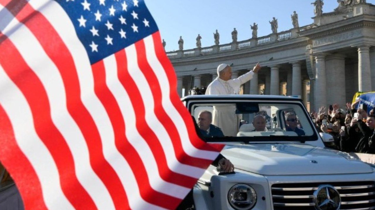 A large US flag fills the left of the frame as Pope Leo XIV, in white, stands in an open white popemobile and waves to a crowd in St. Peter’s Square, with the colonnade and statues behind him under a clear blue sky