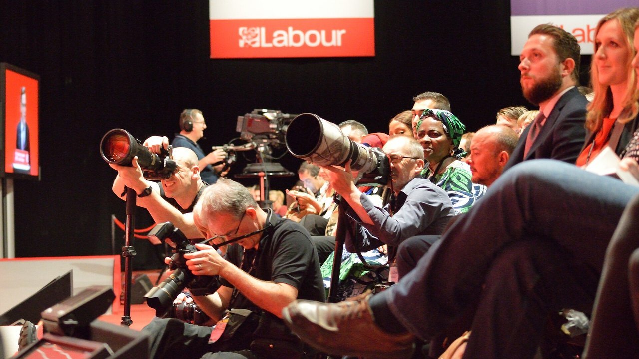 Stand-in, not a photo of this event: a September 2016 file photo of press photographers with long telephoto lenses and a TV camera operator beside a stage under a red Labour sign at a party conference, with audience members seated to the right