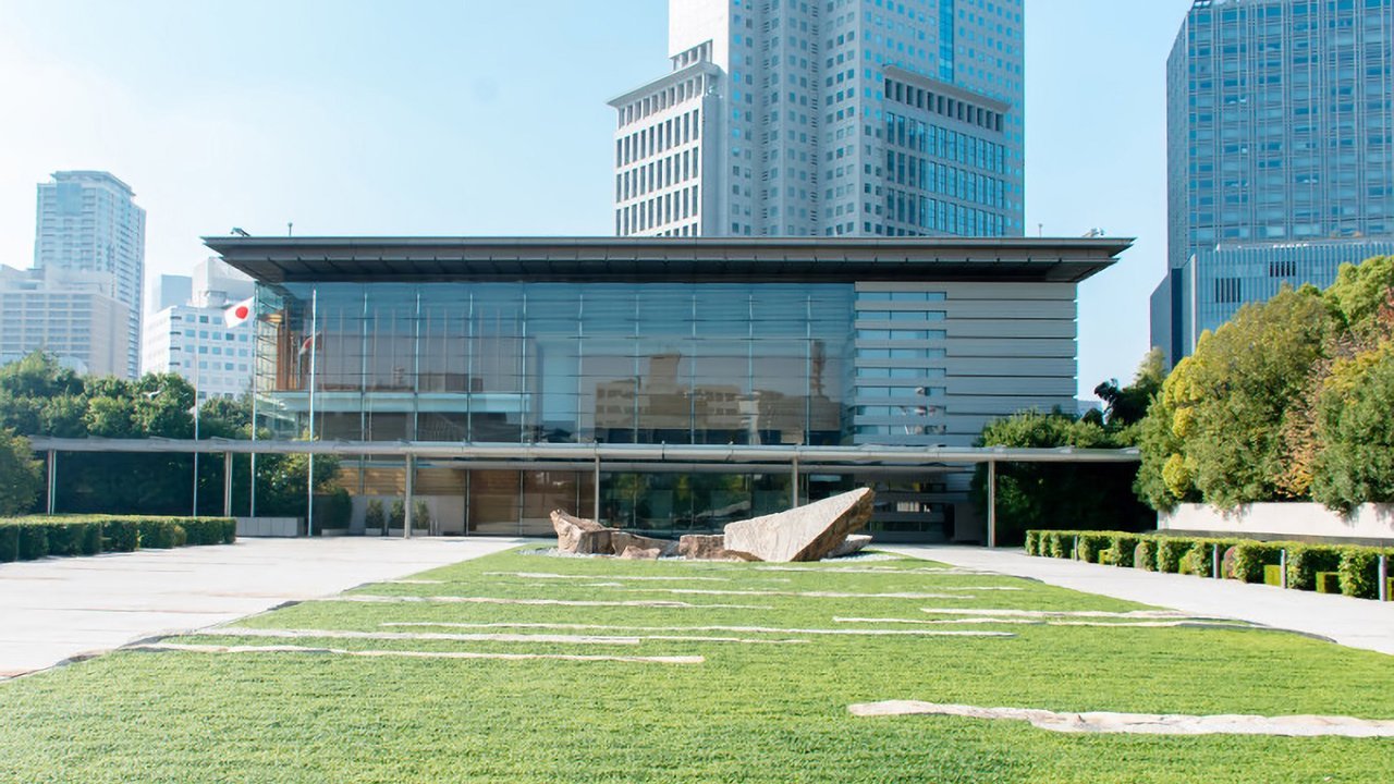 Stand-in, not a photo of this event: a low glass-fronted building with a wide flat roof stands behind a green lawn with stone paths and a rock arrangement, a Japanese flag on a pole at left and high-rise towers behind, at the Prime Minister’s Office in Tokyo around 2020