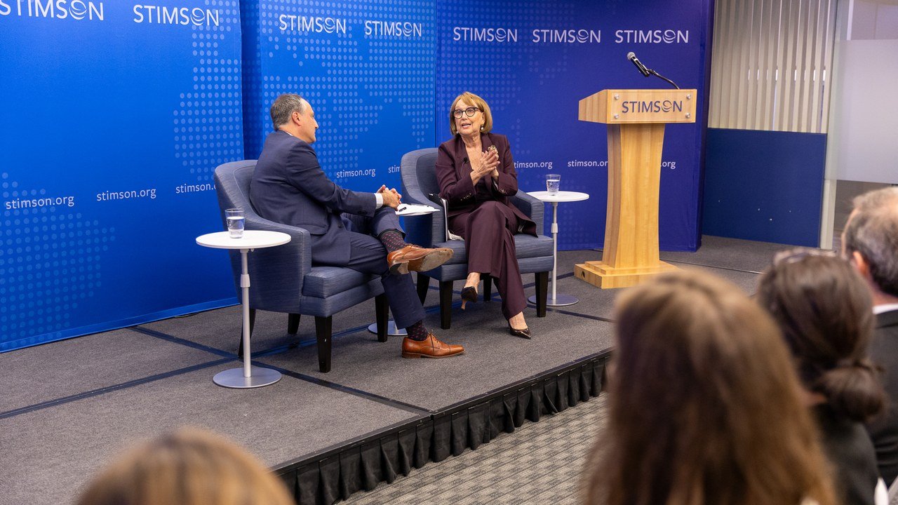 On a stage in front of a blue Stimson Center backdrop, Rebeca Grynspan, in a burgundy suit and glasses, gestures as she speaks from an armchair beside a man in a dark suit, while audience members watch from the foreground