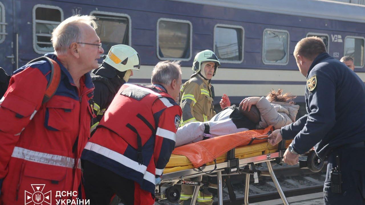 Paramedics in red jackets, firefighters in helmets and a police officer wheel a wounded woman on a stretcher along a Dnipro station platform beside a blue passenger train with shattered windows, in a State Emergency Service photo that Volodymyr Zelenskyy posted on X