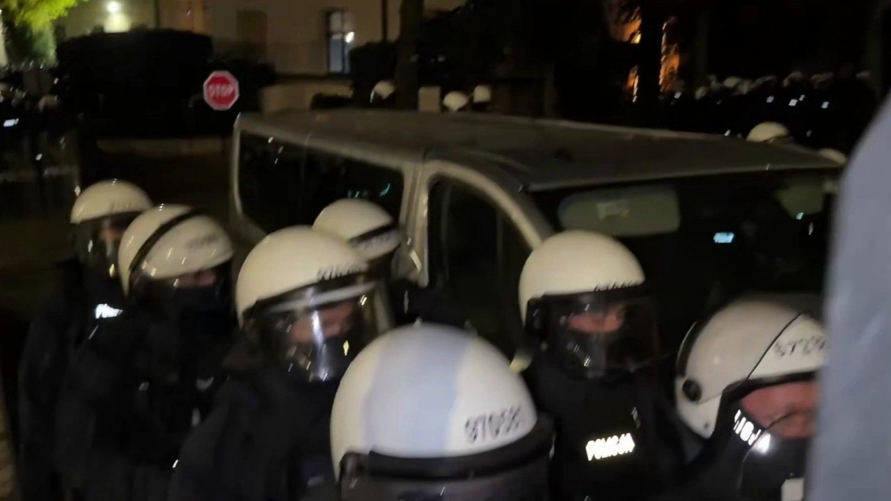 Polish riot police in white helmets with visors down, one vest marked POLICJA, crowd around a silver van on a dark street at night