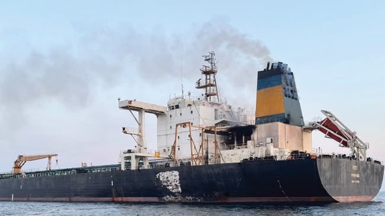A black-hulled tanker with a blue and yellow funnel sits on calm water, with grey smoke drifting from its superstructure, scorch marks on the deckhouse and a blackened patch on the hull, in a photo released by Oman’s Maritime Security Centre