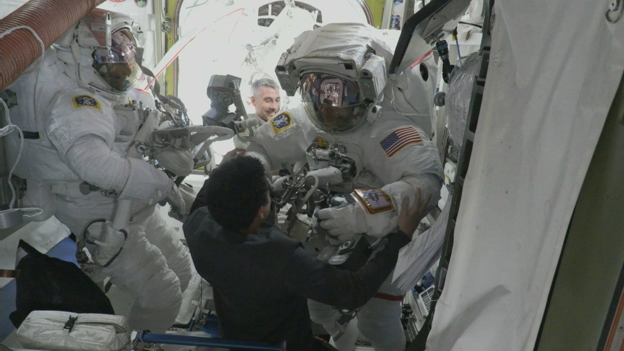 Two astronauts in white spacesuits with helmets on, inside the International Space Station, while two crewmates without suits help them with gear before the Oct. 29 spacewalk, in an image posted on X by Spaceflight Now