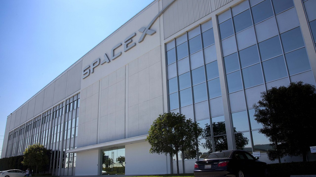 Stand-in, not a photo of this event: the white SpaceX headquarters building in Hawthorne, California, with the SpaceX wordmark on its facade, tall glass windows, trees and a parked dark sedan, in a 2012 company photo