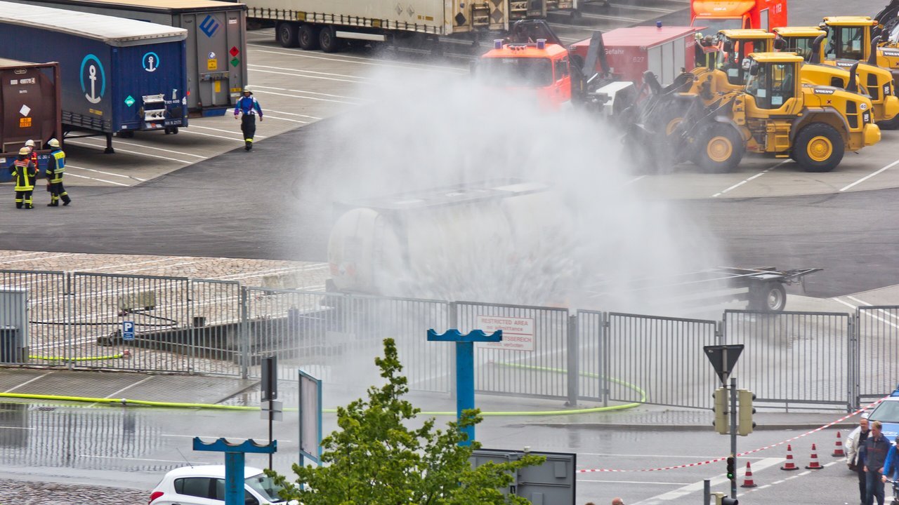 Stand-in, not a photo of this event: at the Schwedenkai ferry terminal in Kiel, Germany, in June 2012, a broad spray of water, which the photographer titled the cooling of a tank container, covers a white cylindrical tank container on a trailer behind a metal fence, while firefighters in helmets and reflective jackets stand at left and red emergency vehicles and yellow wheel loaders wait in the background