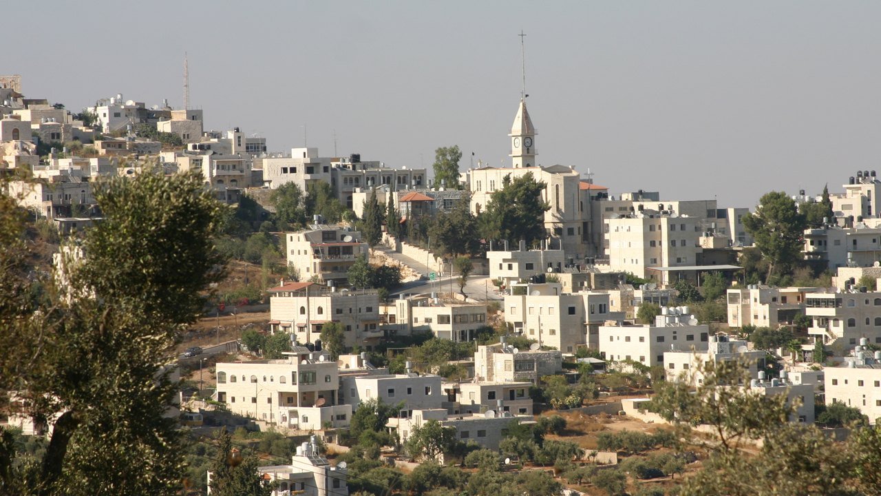 Stand-in, not a photo of this event: hillside village of pale stone houses with a church clock tower near the top and olive trees in the foreground, Taybeh, West Bank, 2010